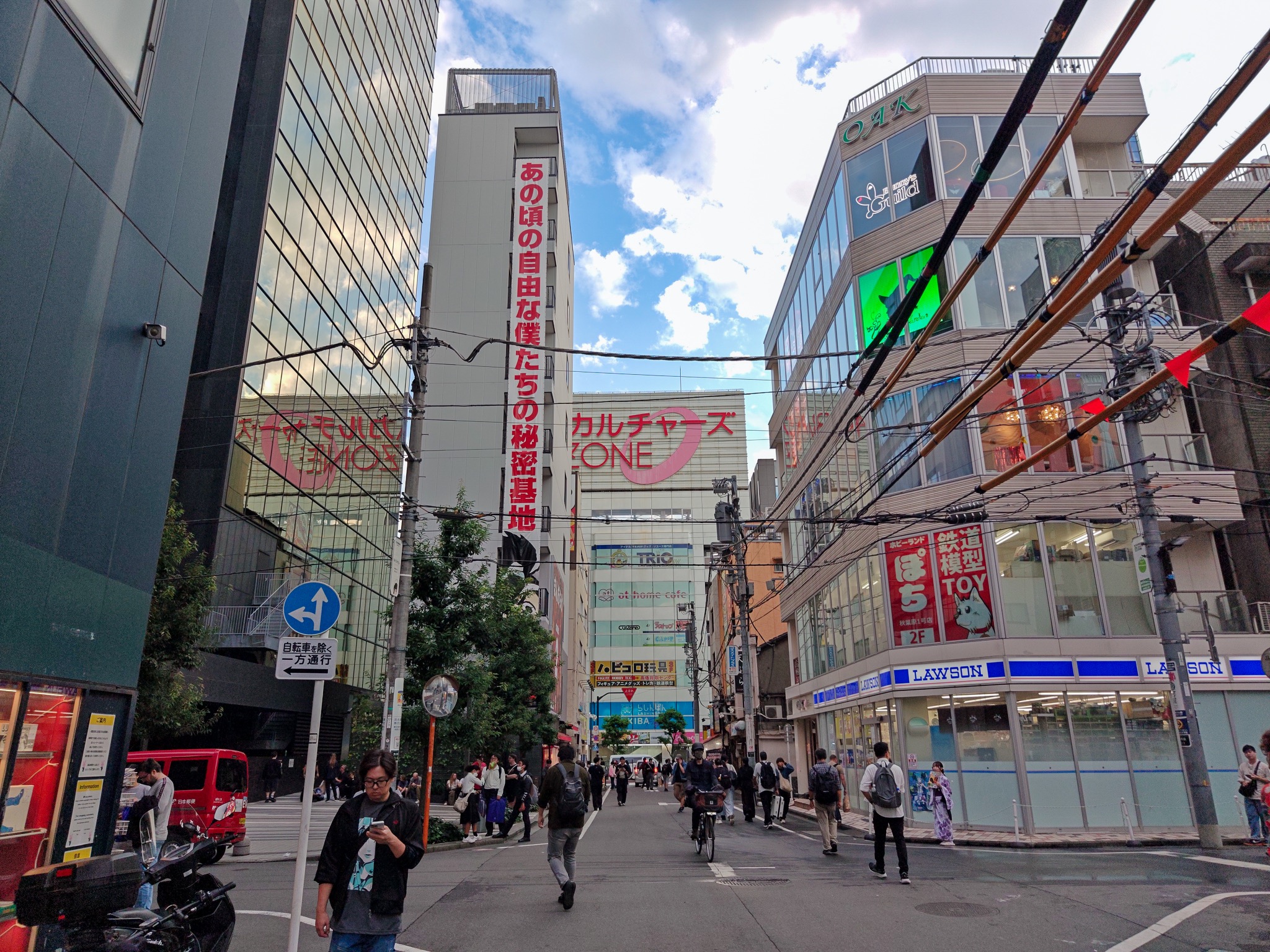 Pedestrians on a street beside Akihabara Cultures Zone and other buildings beneath a blue sky with clouds.