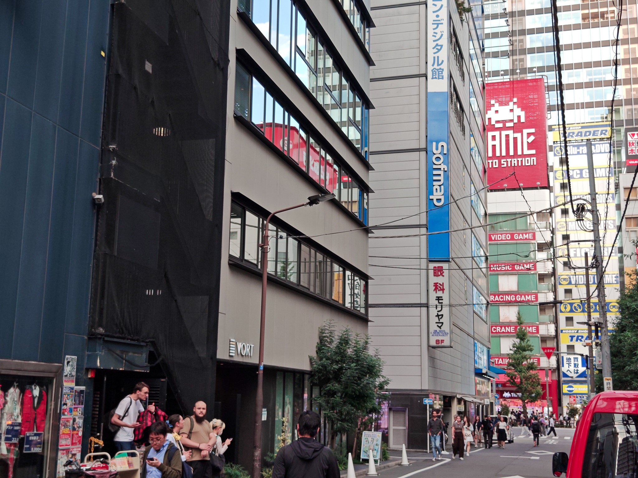 Pedestrians on a street between buildings, with Sofmap, Taito Station, and TRADER signs visible ahead.
